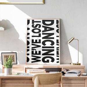 May include: A black and white poster with the text 'DANCING WE'VE LOST' in a modern font. The poster is framed in a light wood frame and is hanging on a white wall above a wooden desk with a gold lamp and a potted plant.