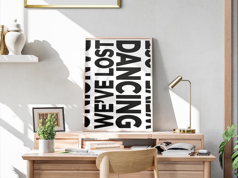May include: A black and white poster with the text 'DANCING WE'VE LOST' in a modern font. The poster is framed in a light wood frame and is hanging on a white wall above a wooden desk with a gold lamp and a potted plant.