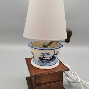 Könnte beinhalten: Eine Vintage-Lampe mit weißem Lampenschirm, blau-weißem Keramiksockel mit Windmühlendesign und Holzsockel mit Schublade. Die Lampe hat einen dunkelbraunen Holzgriff und ein weißes Netzkabel.