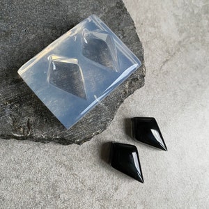 Op de afbeelding: Twee zwarte, ruitvormige oorbellen van hars. De oorbellen worden getoond naast een heldere siliconen mal met twee ruitvormen.