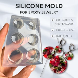 Könnte beinhalten: Transparente Silikonform zum Herstellen von Epoxidharzschmuck. Die Form hat sechs runde Vertiefungen. Die Form wird in einer Hand gehalten. Es gibt drei fertige Epoxidharzstücke mit floralen Designs auf weißem Hintergrund.