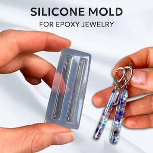 Peut inclure: Un moule en silicone transparent pour la fabrication de bijoux en résine époxy. Le moule comporte deux cavités rectangulaires. Une paire de longues boucles d'oreilles rectangulaires avec un motif floral est présentée à côté du moule.