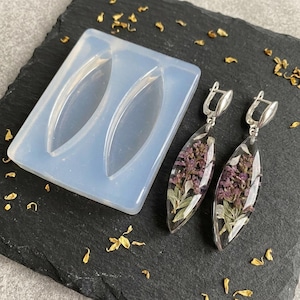 May include: A translucent silicone mold with two marquise-shaped cavities alongside a pair of matching drop earrings. Each earring features a clear resin marquise pendant containing dried purple flowers and foliage, attached to silver-toned metal hooks.