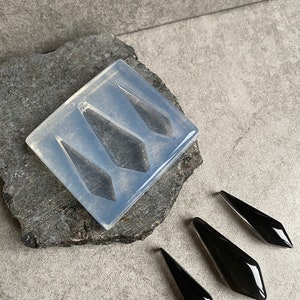 Op de afbeelding: Een transparante siliconen mal met vier diamantvormige holtes. Naast de mal zijn drie zwarte, diamantvormige oorbellen van hars te zien.