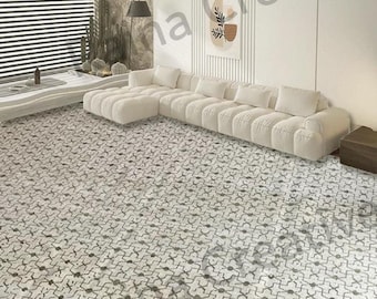 May include: A large area rug featuring a repeating geometric pattern in shades of grey and white. The rug covers the floor of a room containing a long, white, tufted sectional sofa, a wall-mounted abstract art piece, and a window with blinds.