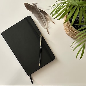 Może przedstawiać: Czarny notes, oprawiony w skórę, z długopisem w kolorze złotym i piórem na białym tle.
