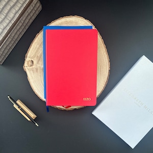 Può includere: Due quaderni, uno rosso e uno blu, sono impilati su una fetta di legno. Il quaderno rosso ha la scritta "EIZO" stampata in oro sulla copertina. Una scatola bianca con il testo "ELIZABETH MILAN LONDON" si trova nell'angolo in basso a destra dell'immagine.