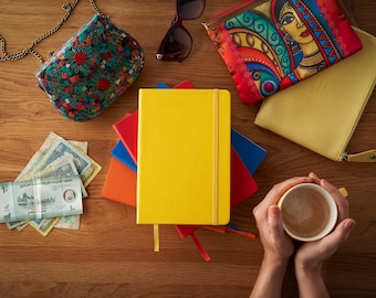 May include: A yellow notebook with a red, orange, blue, and black cover sits on a wooden table with a cup of coffee, a purse, and money.