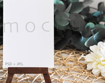May include: A white mockup card on a small wooden easel, with the word "mock" partially visible. The card is set against a backdrop of greenery and a woven mat, with the text "PSD + JPG" at the bottom.
