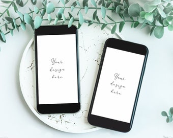 May include: Two black smartphones with blank white screens are lying on a white plate with a white background. Green leafy branches are on either side of the plate.
