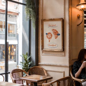 K&ouml;nnte beinhalten: Ein gerahmter Druck mit wei&szlig;em Hintergrund und goldenem Rahmen. Der Druck zeigt eine stilisierte Illustration eines Croissants und einer Tasse Kaffee mit dem Text "But first, breakfast".