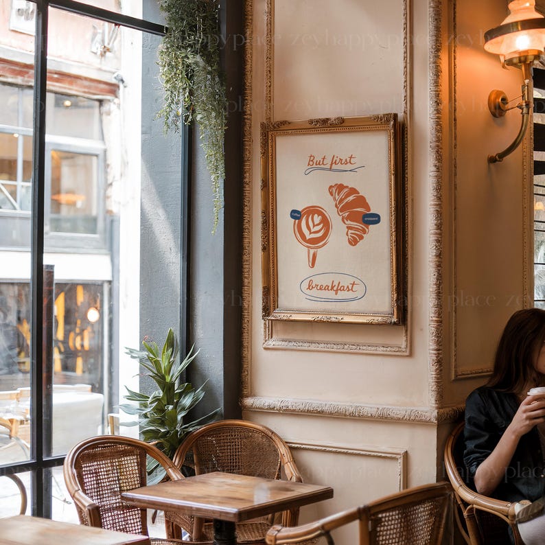 K&ouml;nnte beinhalten: Ein gerahmter Druck mit wei&szlig;em Hintergrund und goldenem Rahmen. Der Druck zeigt eine stilisierte Illustration eines Croissants und einer Tasse Kaffee mit dem Text "But first, breakfast".