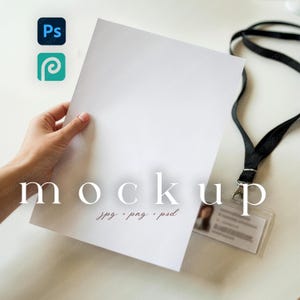 May include: A white paper mockup held by a hand, with the word "mockup" in a serif font overlaid on the image. The image also includes a lanyard with an ID badge and Adobe Photoshop and Adobe Illustrator icons.