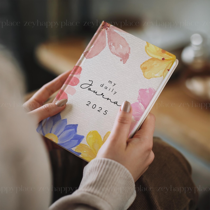Puede incluir: Un diario blanco con un dise&ntilde;o floral. La portada presenta flores de acuarela en tonos rosa, amarillo y azul. El texto "my daily journal 2025" est&aacute; impreso en negro en la portada.