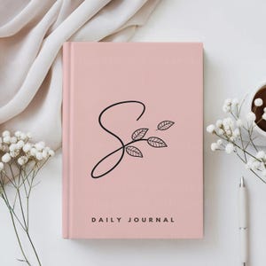 K&ouml;nnte beinhalten: Ein rosa t&auml;gliches Tagebuch mit einem schwarzen stilisierten Buchstaben S und einem kleinen Zweig mit Bl&auml;ttern. Der Text "DAILY JOURNAL" ist in Schwarz am unteren Rand des Deckels gedruckt.