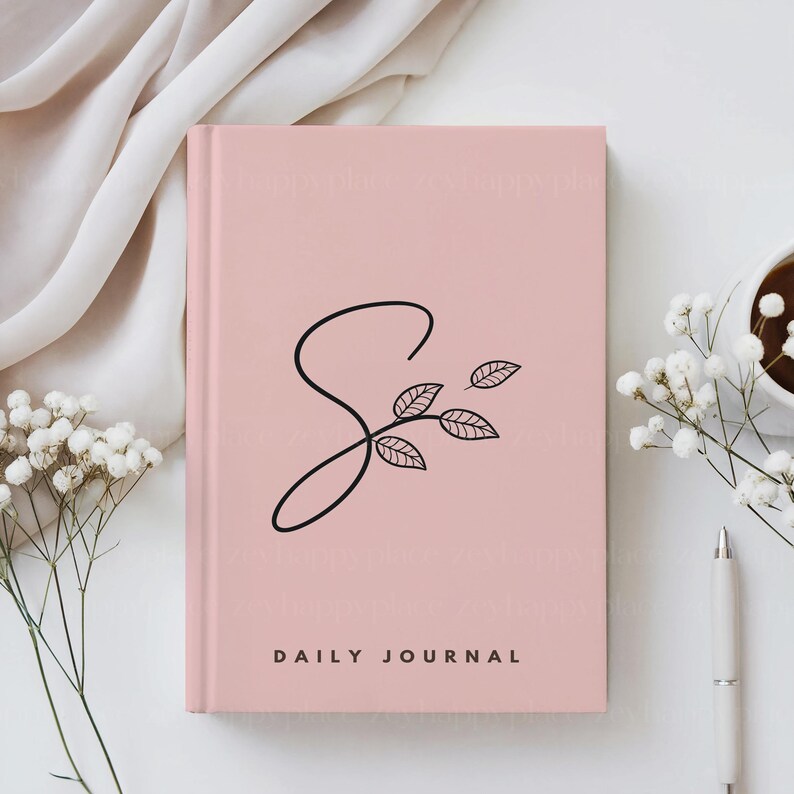 K&ouml;nnte beinhalten: Ein rosa t&auml;gliches Tagebuch mit einem schwarzen stilisierten Buchstaben S und einem kleinen Zweig mit Bl&auml;ttern. Der Text "DAILY JOURNAL" ist in Schwarz am unteren Rand des Deckels gedruckt.
