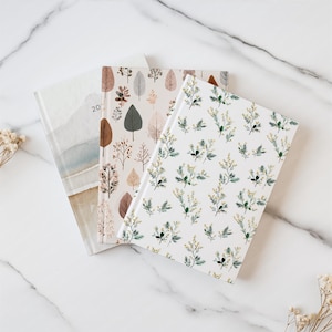 May include: Three notebooks with floral and leaf patterns. One notebook has a watercolor design with the number "20" visible. The other two notebooks feature botanical designs in shades of brown, green, and white. The notebooks are arranged on a white marble surface.