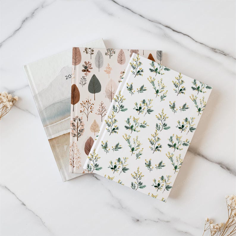 May include: Three notebooks with floral and leaf patterns. One notebook has a watercolor design with the number "20" visible. The other two notebooks feature botanical designs in shades of brown, green, and white. The notebooks are arranged on a white marble surface.