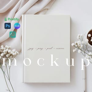 May include: White blank notebook mockup with a white pen, white flowers, and a white fabric in the background. The text "jpg - png - psd - canva mockup" is written in pink on the notebook.