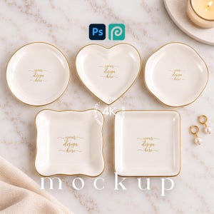 May include: Five white ceramic trinket dishes in various shapes including round, heart, wavy square, and square, each featuring a gold rim and the gold text your design here printed in the center, arranged on a light marble surface.