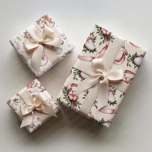 May include: Three wrapped gifts with cream-colored bows. The wrapping paper features floral and unicorn patterns in pink and cream. The gifts are arranged on a white surface, creating a visually appealing presentation.