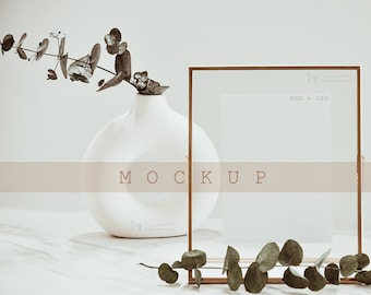 May include: A gold-toned frame mockup with a white background. The frame is sitting on a white surface with a vase of eucalyptus branches to the left. The text "MOCKUP" is printed in brown on a light brown band across the middle of the image.