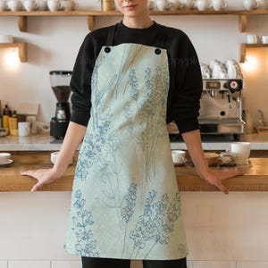 May include: A light blue apron with a floral pattern of lavender flowers. The apron has two adjustable straps and a pocket.