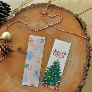 Puede incluir: Dos marcap&aacute;ginas con tem&aacute;tica navide&ntilde;a. Un marcap&aacute;ginas presenta un fondo rosa y azul con hombres de jengibre, bastones de caramelo, copos de nieve y regalos. El otro marcap&aacute;ginas presenta un &aacute;rbol de Navidad verde con adornos rojos y blancos, bastones de caramelo y una pancarta roja con el texto "Feliz Navidad".
