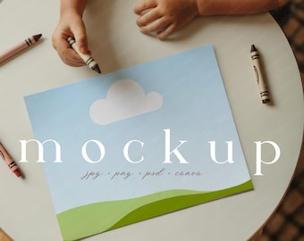 Op de afbeelding: Een mockup-ontwerp met een lichtblauwe lucht, een witte wolk en groene heuvels. Het woord "mockup" staat in het wit, met "jpg, png, psd, canva" eronder. Kleurpotloden liggen verspreid rond het papier. Het ontwerp bevat de logo's voor Photoshop, Canva en een derde app.