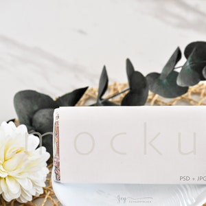 May include: White rectangular box mockup with the word "mockup" in gray text. The box is sitting on a white plate with a white background. There are green leaves and a white flower in the background.