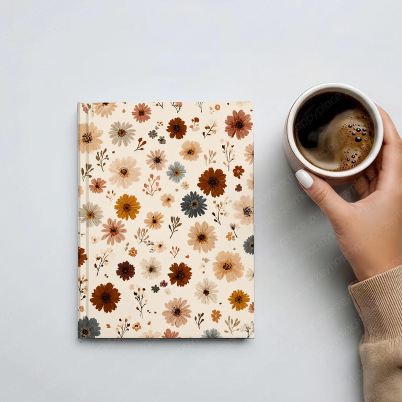 May include: A floral-patterned journal with a cream-colored background and various flowers in shades of brown, yellow, and blue. A cup of coffee is held in the hand next to the journal.