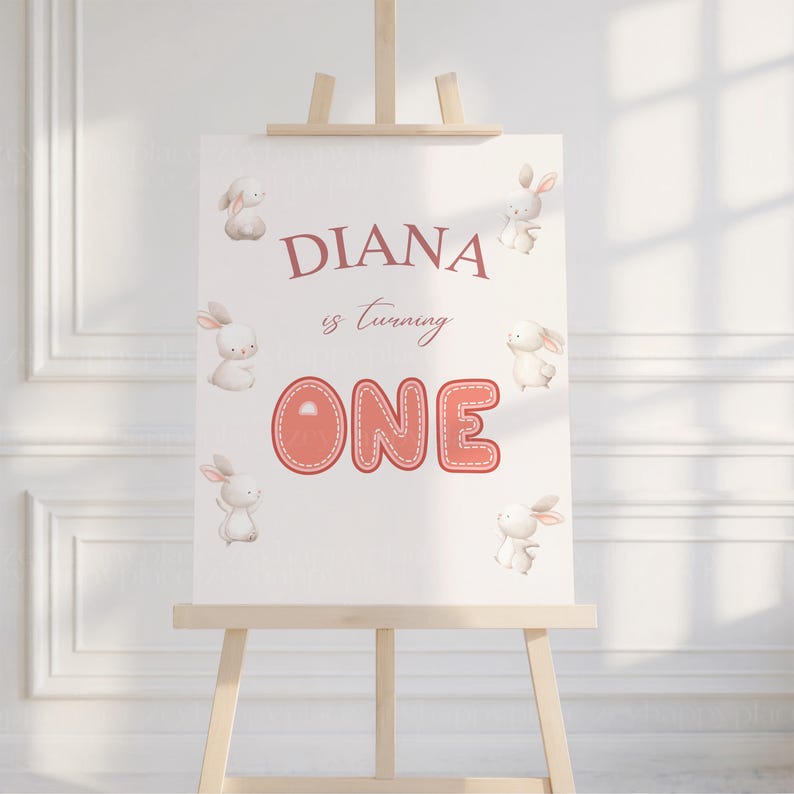 May include: A white sign with pink text and bunny illustrations. The sign says "Diana is turning one".