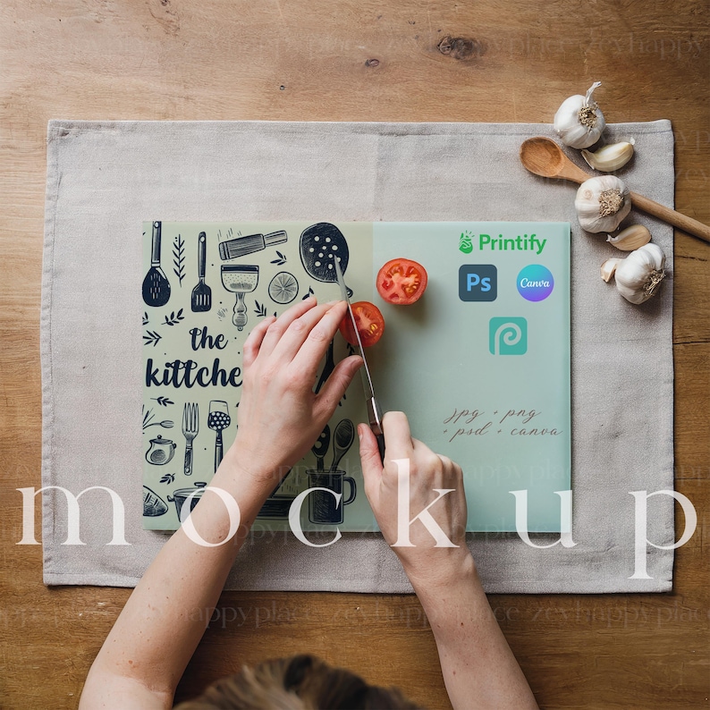 Op de afbeelding: Een persoon snijdt een tomaat op een groen snijplank met een zwart-wit ontwerp met de tekst "the kitche". Het snijplank ligt op een beige linnen placemat. Er liggen twee rode tomaten en een paar teentjes knoflook op het snijplank.