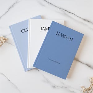 May include: Three hardcover journals in shades of blue and white are arranged on a marble surface. The journals are labeled with names: "OLIVIA", "JAMES", and "HANNAH". The blue journal reads "ALL THAT WENT UNSAID".