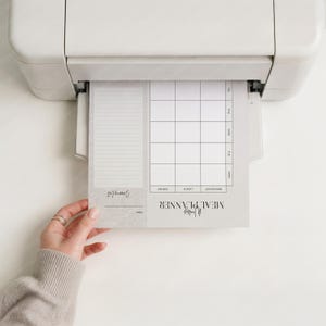 May include: A weekly meal planner being printed from a white printer. The planner has a section for a shopping list and a grid for daily meal planning. The text "Weekly Meal Planner" is visible.