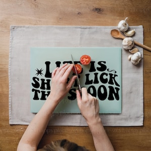 Op de afbeelding: Een lichtblauw snijplank met de tekst "Life is short, eat the cake soon" in zwart. Een persoon snijdt een rode tomaat op het plank met een zilveren mes.
