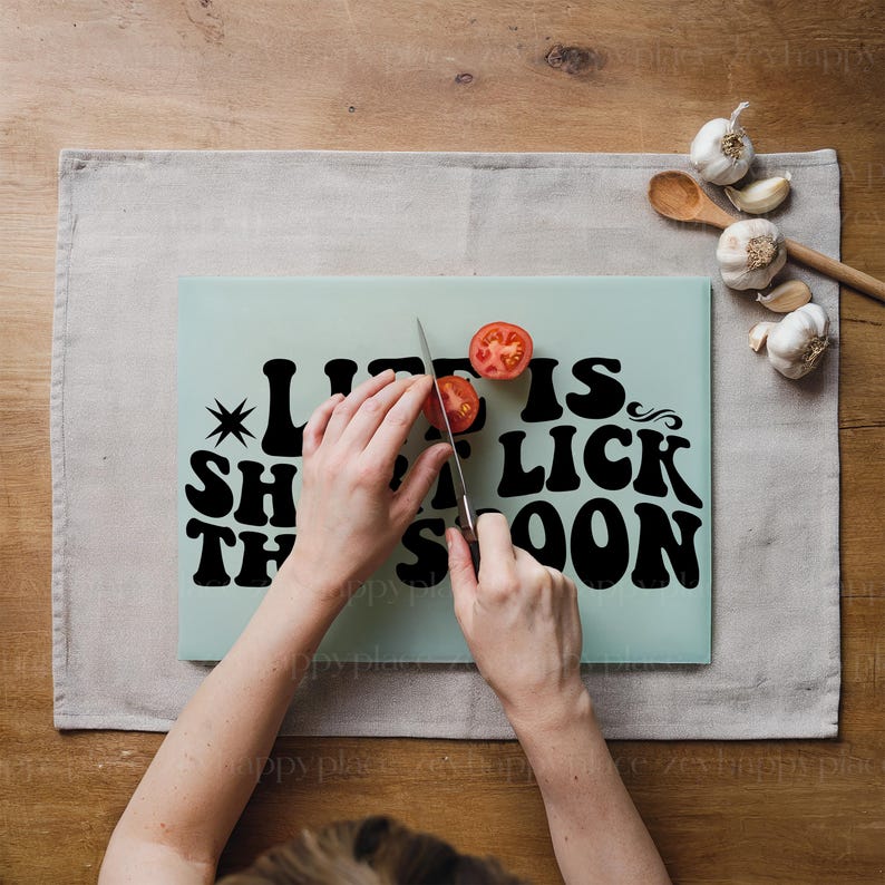 Op de afbeelding: Een lichtblauw snijplank met de tekst "Life is short, eat the cake soon" in zwart. Een persoon snijdt een rode tomaat op het plank met een zilveren mes.