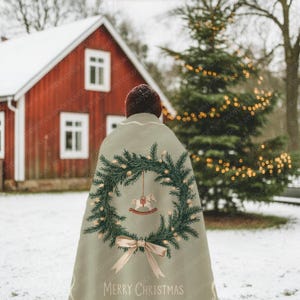 May include: A light green Christmas cape with a festive design. The cape features a wreath of evergreen branches, fairy lights, and a rocking horse ornament. The words "MERRY CHRISTMAS" are printed at the bottom, with a light brown bow.