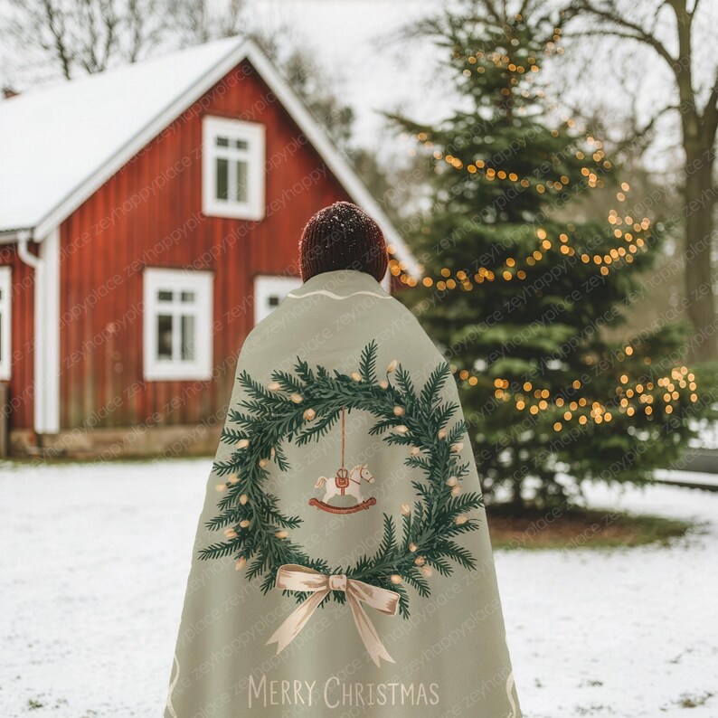 May include: A light green Christmas cape with a festive design. The cape features a wreath of evergreen branches, fairy lights, and a rocking horse ornament. The words "MERRY CHRISTMAS" are printed at the bottom, with a light brown bow.