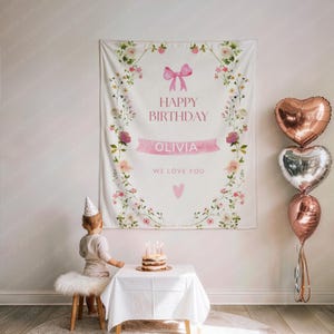May include: A white birthday banner with floral and pink bow accents. The banner reads "HAPPY BIRTHDAY OLIVIA WE LOVE YOU." A small cake with lit candles sits on a table. Heart-shaped balloons are to the right.