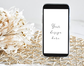 May include: A black smartphone with a white screen displaying the text "Your design here" is sitting on a woven mat with a white flower arrangement and a white ribbon.