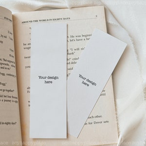 K&ouml;nnte beinhalten: Zwei leere wei&szlig;e Lesezeichen mit dem Text "Your design here" darauf gedruckt. Die Lesezeichen liegen auf einem ge&ouml;ffneten Buch mit Text &uuml;ber eine Reise um die Welt in achtzig Tagen.