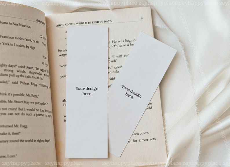 K&ouml;nnte beinhalten: Zwei leere wei&szlig;e Lesezeichen mit dem Text "Your design here" darauf gedruckt. Die Lesezeichen liegen auf einem ge&ouml;ffneten Buch mit Text &uuml;ber eine Reise um die Welt in achtzig Tagen.