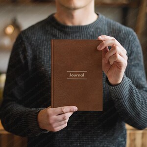 May include: A brown journal with the word "Journal" printed in white. The journal is held by a person wearing a gray sweater. The journal has a textured cover.