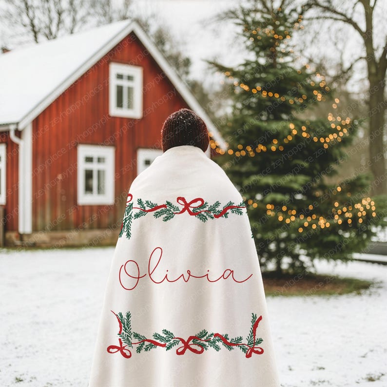 May include: A cream-colored blanket with red and green Christmas garland embroidery and the name "Olivia" in red script. The blanket is draped over a person standing in a snowy outdoor scene with a red house and Christmas tree.