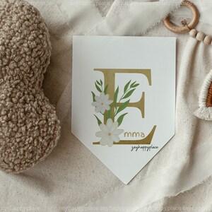 May include: A white banner featuring a gold letter E adorned with white flowers and green leaves. The banner includes the text "mma" and "zeyhappyplace" printed on it.