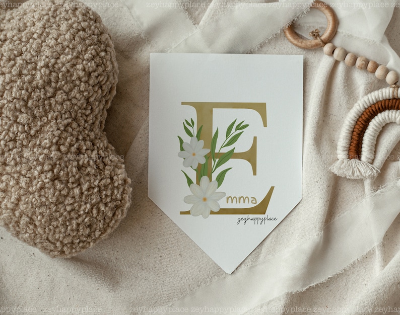 May include: A white banner featuring a gold letter E adorned with white flowers and green leaves. The banner includes the text "mma" and "zeyhappyplace" printed on it.