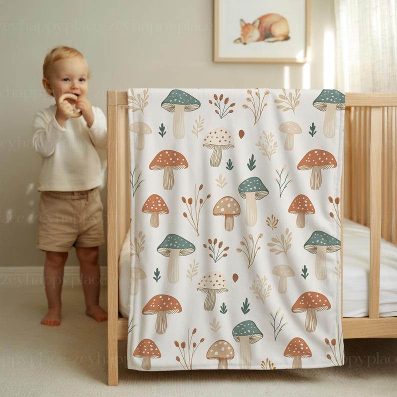 May include: A white baby blanket featuring a repeating pattern of mushrooms in brown, teal, and beige, with sprigs and leaves. The blanket is draped over a wooden cot.