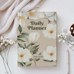 K&ouml;nnte beinhalten: Ein beiger Tagesplaner mit einem floralen Design mit wei&szlig;en Blumen und gr&uuml;nen Bl&auml;ttern. Der Text "Daily Planner" ist in Gr&uuml;n auf dem vorderen Umschlag gedruckt. Der Text "hello 2025!" ist in Gr&uuml;n am unteren Rand des Umschlags gedruckt.