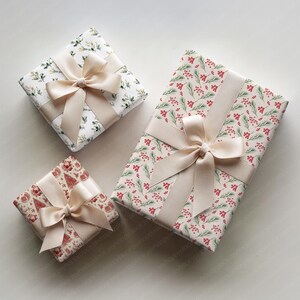 May include: Three wrapped gifts with cream-colored bows. The wrapping paper features festive patterns: one with green leaves, another with red berries, and a third with Christmas tree designs. The gifts are arranged on a white surface.